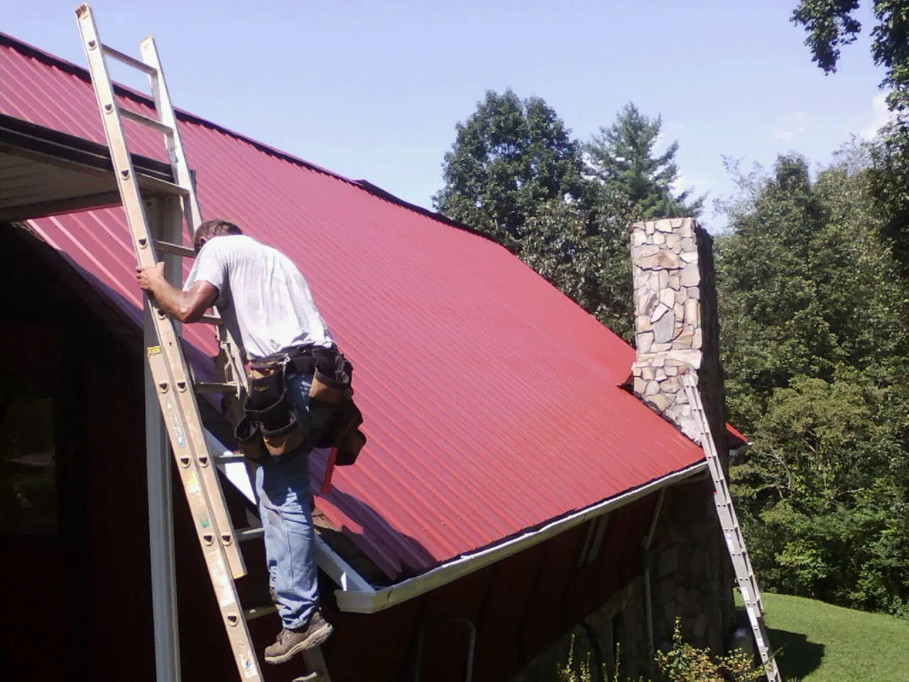 Roofer on a ladder beside a red metal roof and stone chimney — Soffit & Fascia Repair by Roofing Brothers in Sawyer